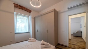 A bedroom with a bed, two folded towels, a wardrobe, a window with blinds, and an air conditioning unit. - Villa Cadiz Apartments Varazze (Varazze)