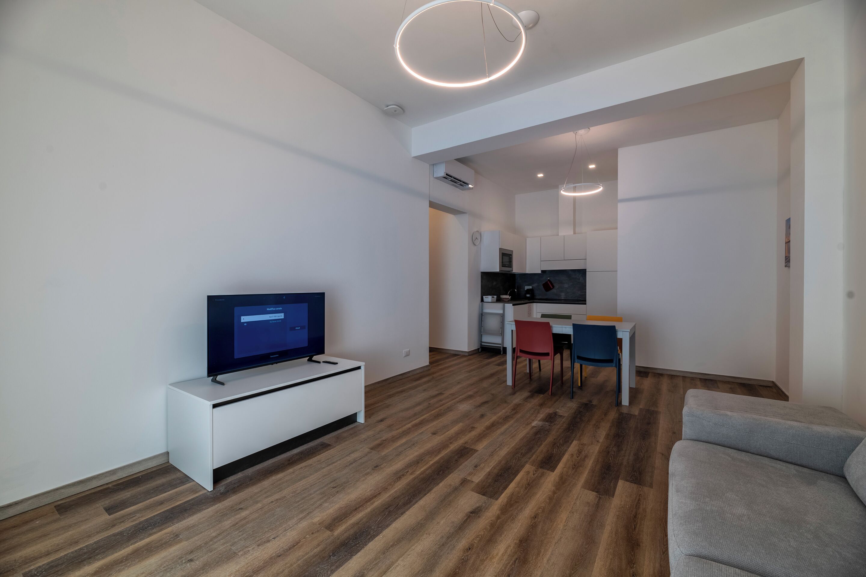Una sala de estar moderna con un televisor de pantalla plana sobre una consola blanca, un sofá gris y una zona de comedor con una mesa y sillas.