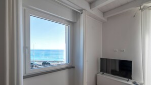 A room with a large window overlooking the sea, a flat-screen TV mounted on the wall, and a wooden floor. - Villa Cadiz Apartments Varazze (Varazze)