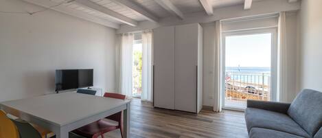 En rummelig stue med trægulv, hvidt loft, grå sofa, spisebord med farverige stole, tv og en skydedør til en balkon med havudsigt.