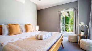 A hotel room with a large bed, a small table with a plant, and a view of greenery through the window. - Villa Madison | Port Douglas (PORT DOUGLAS)