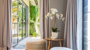 A room with a large window, a potted orchid, a woven ottoman, and a small table with a bowl. - Villa Madison | Port Douglas (PORT DOUGLAS)