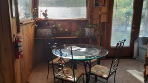 Un comedor con una mesa redonda de vidrio y sillas de metal, un extintor en la pared y una ventana con persianas.