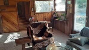 Interior acogedor de una cabaña con techo de madera, una mecedora, una mesa de vidrio y un sofá con cojines.