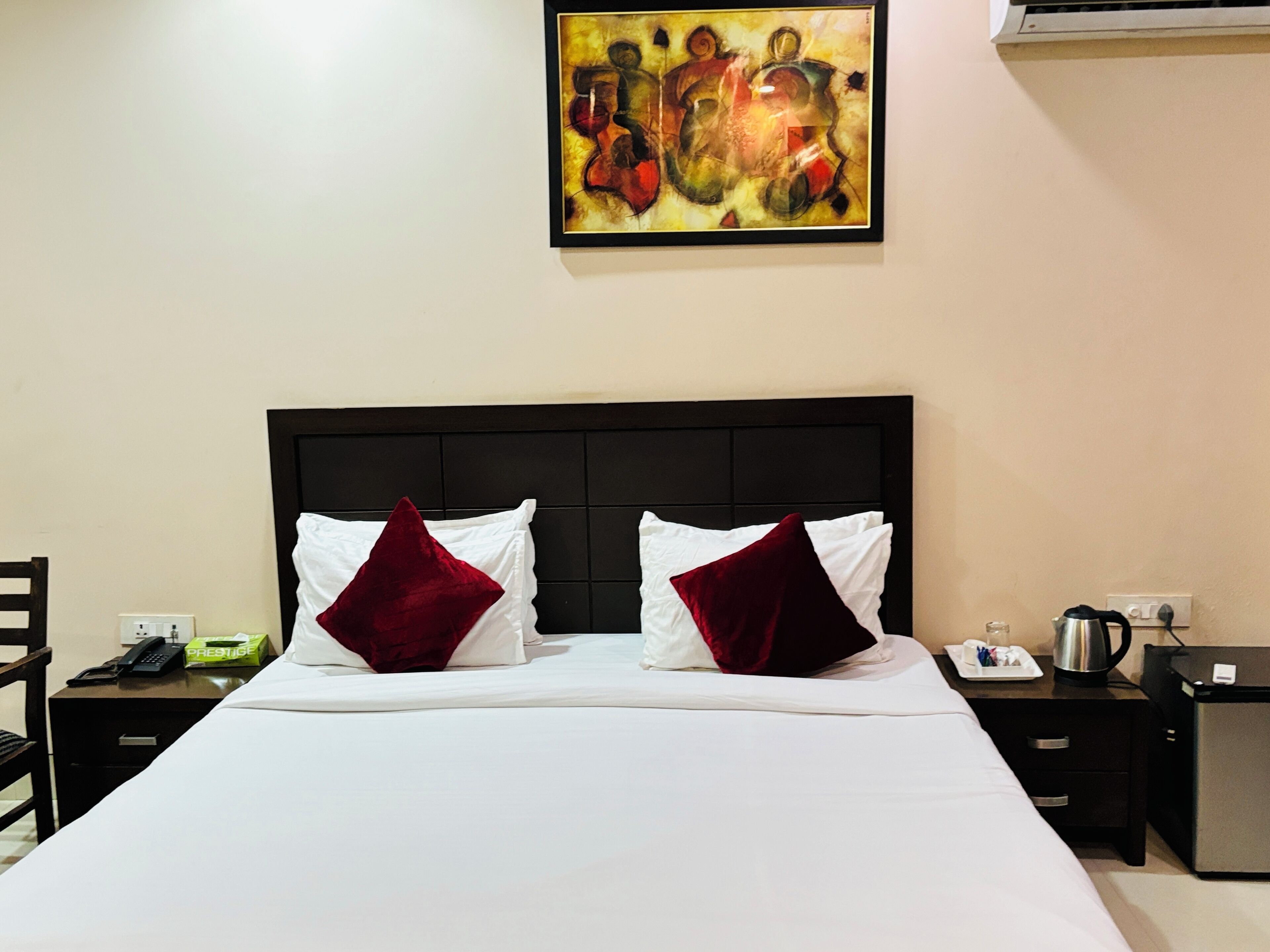 A hotel room with a large bed, two pillows, a nightstand with a phone and a kettle, and a painting on the wall.