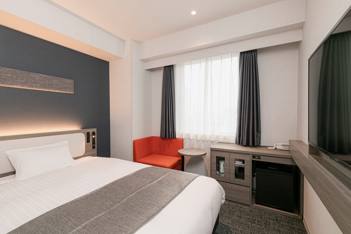 A hotel room with a large bed, a desk with a computer, a chair, a small table, a TV, and a window with curtains.