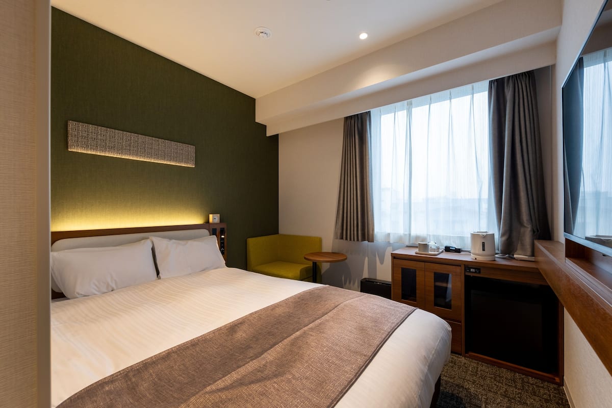 A hotel room with a large bed, a desk with a chair, a small table, and a window with curtains.