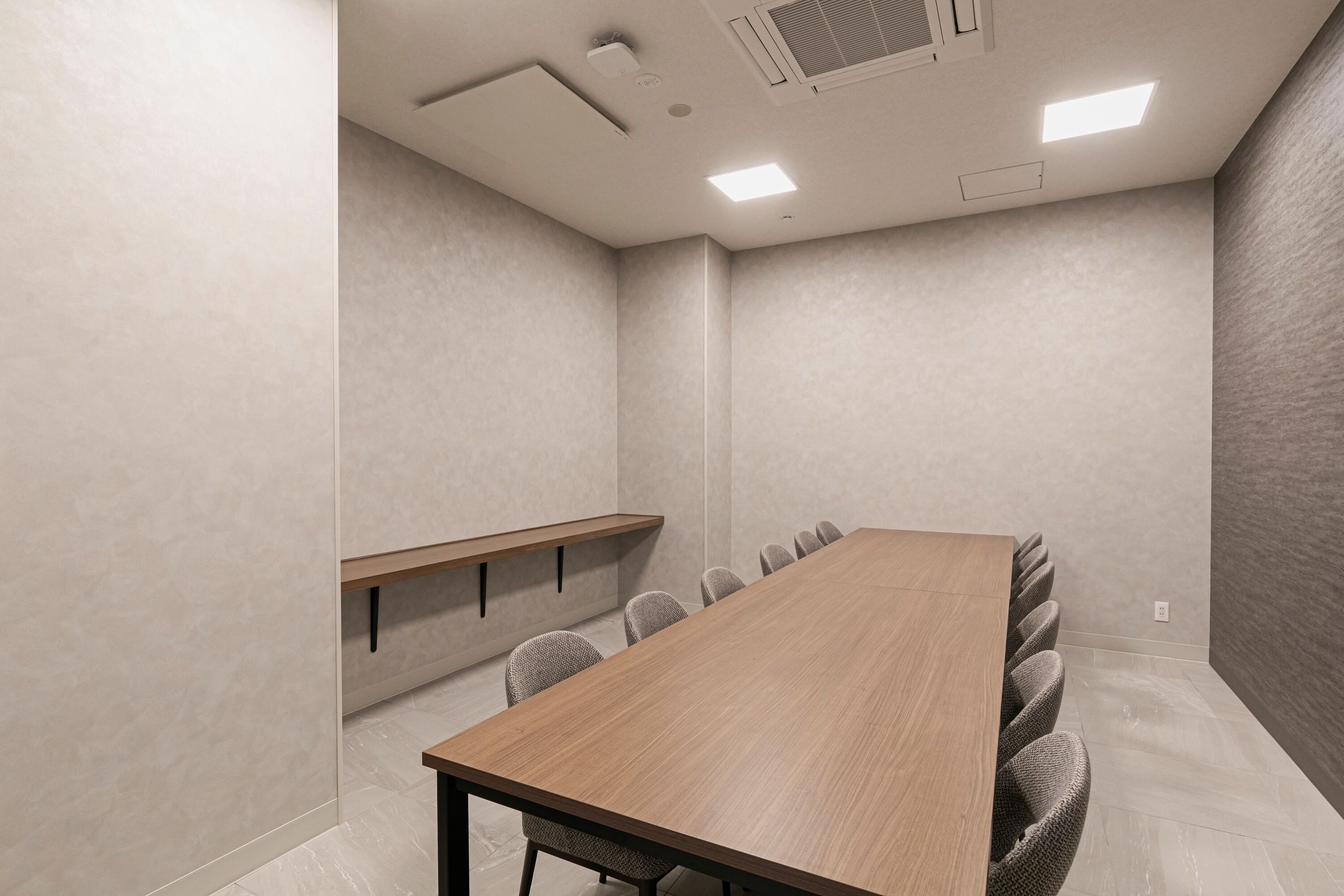 A conference room with a long wooden table, grey upholstered chairs, a small shelf, and recessed ceiling lights.