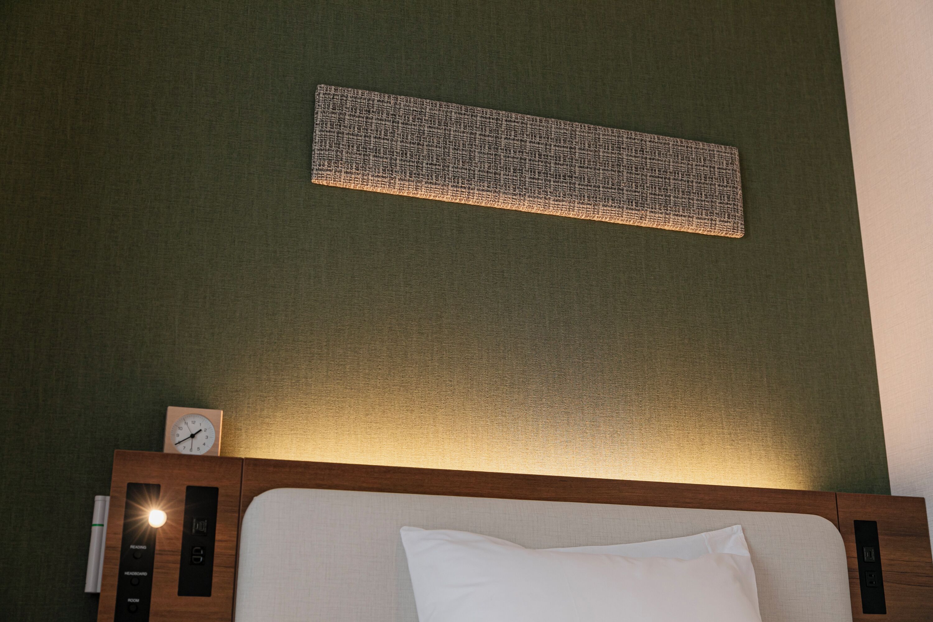 A hotel room with a wooden headboard, a white pillow, a wall clock, and a textured wall panel.