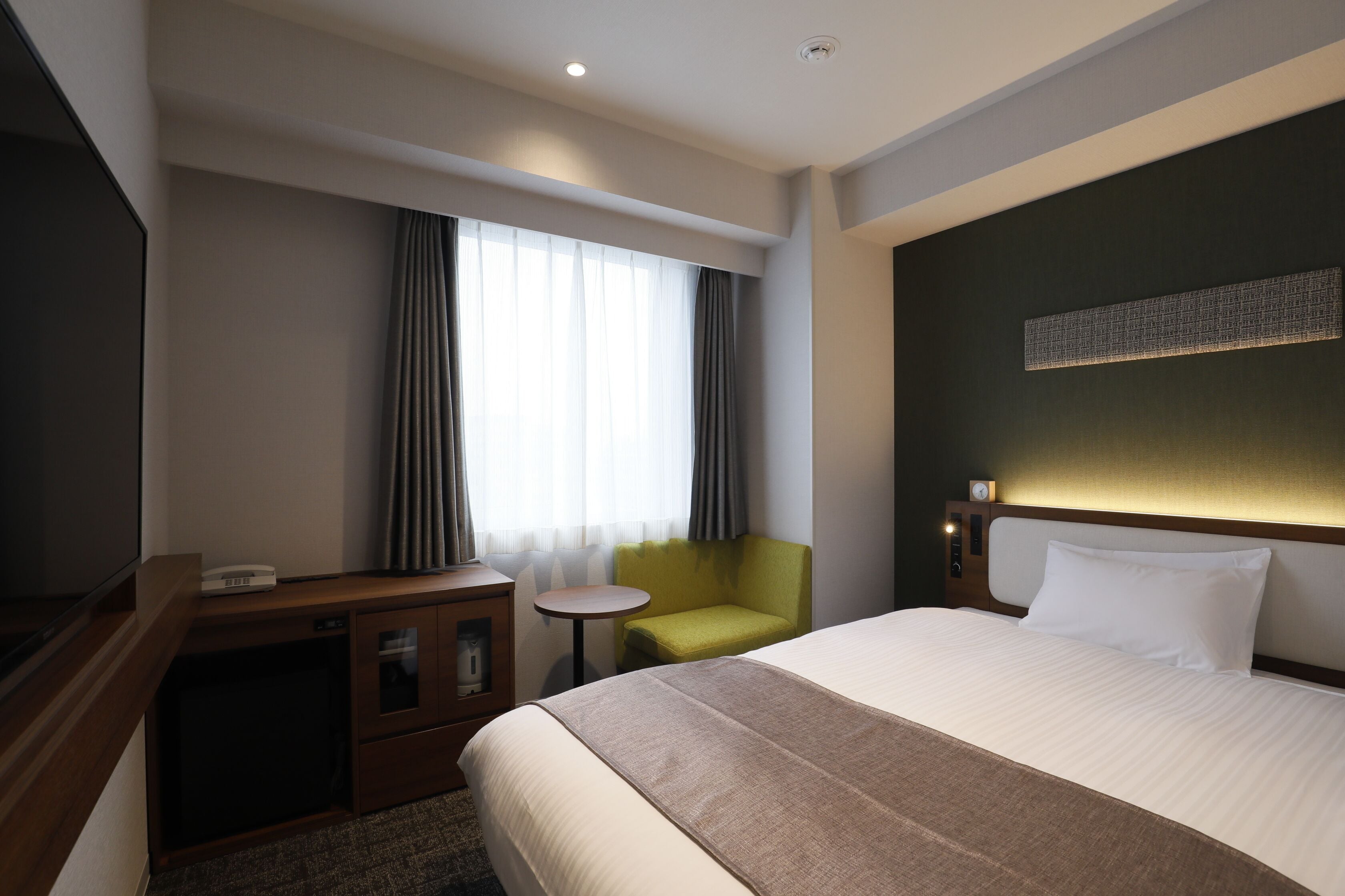 A hotel room with a bed, a desk with a lamp, a small table, and a chair.