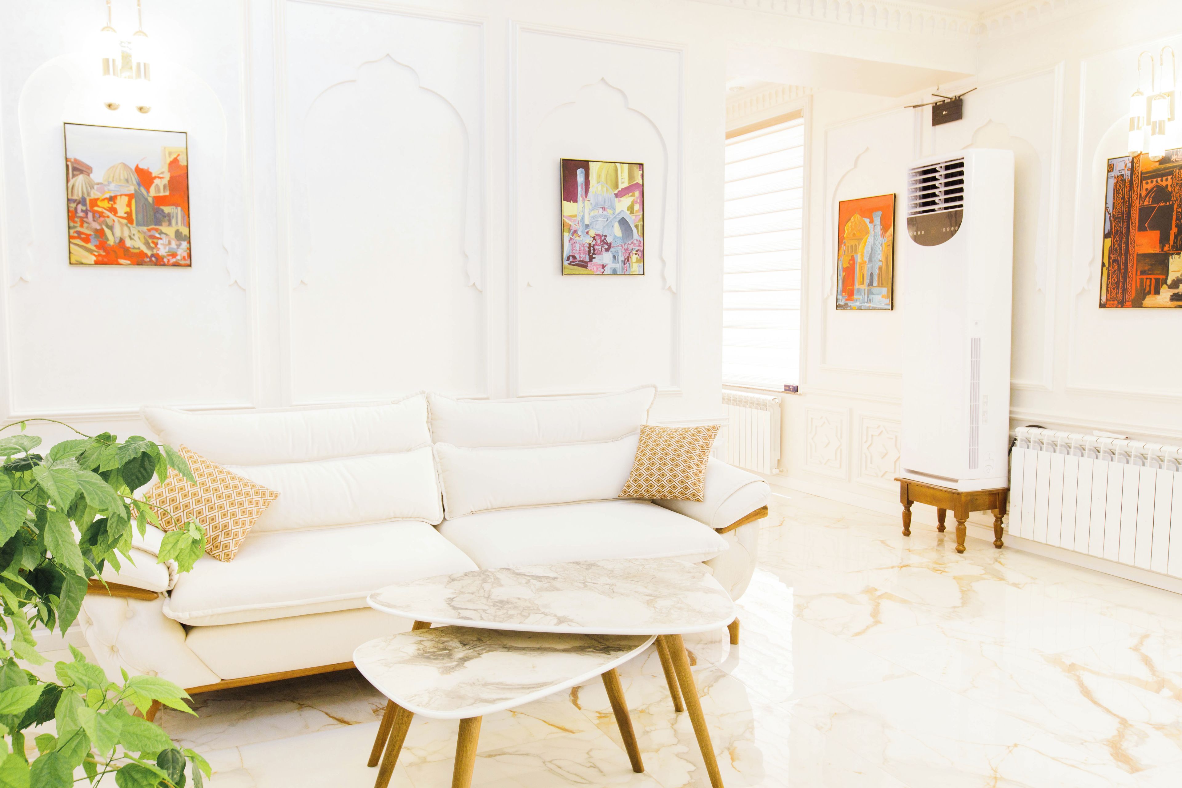 A white sofa with patterned cushions, a marble coffee table, and a radiator in a bright room with artwork on the walls.