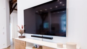 A modern living room with a large flat-screen TV mounted on a white wall, wooden cabinets below, and a wooden floor. - Gute Auszeit - Ferienwohnung Lichtblick (Borken)
