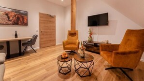 A modern living room with a wooden floor, two armchairs, a coffee table, a TV mounted on the wall, and a wooden door. - Hof Gerwing - Appartement 4, 1-4 Pers (Ahaus-Alstätte)