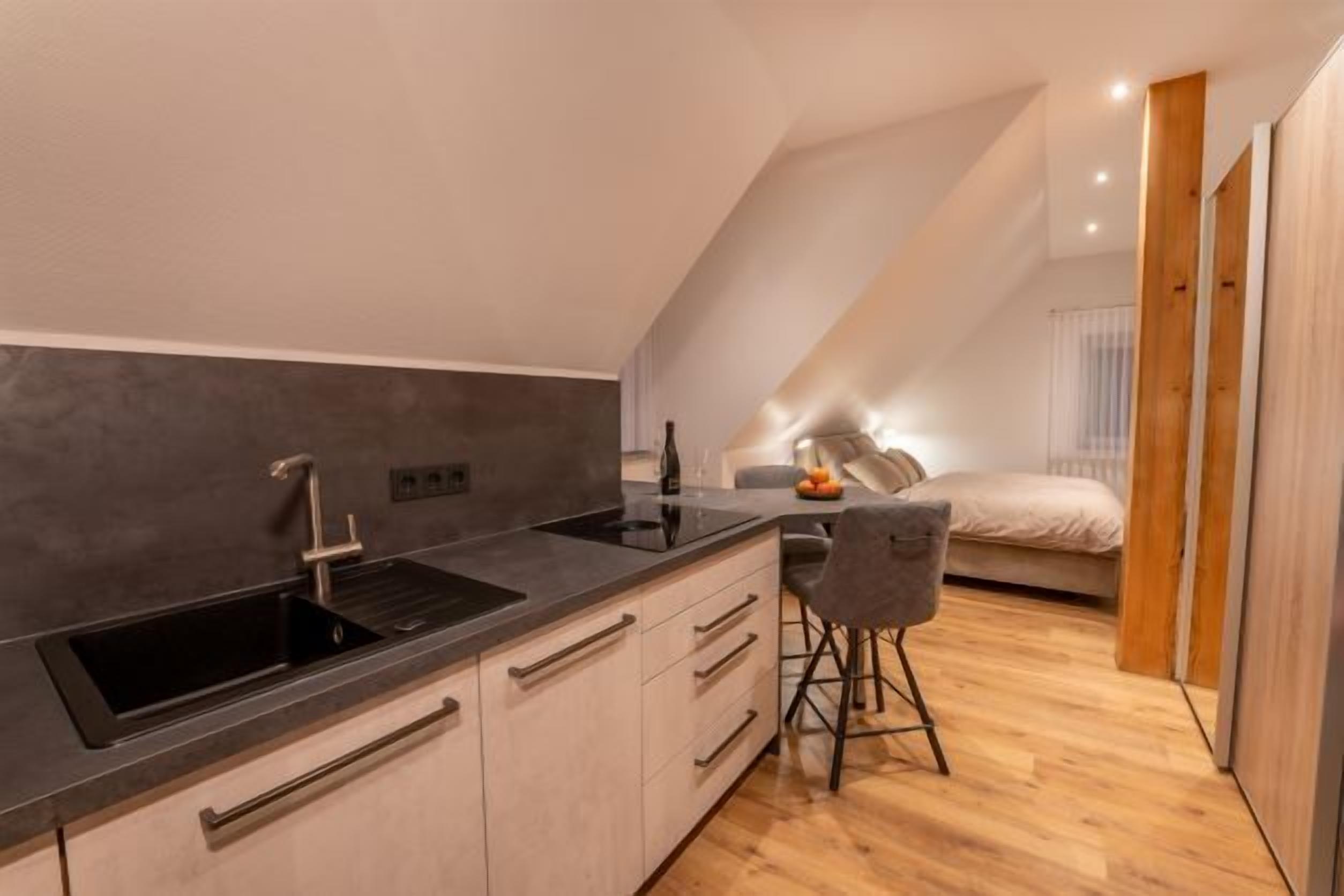 Una cocina moderna con encimera negra, pileta de acero inoxidable y muebles de madera. Hay un taburete y una cama en la habitación contigua.