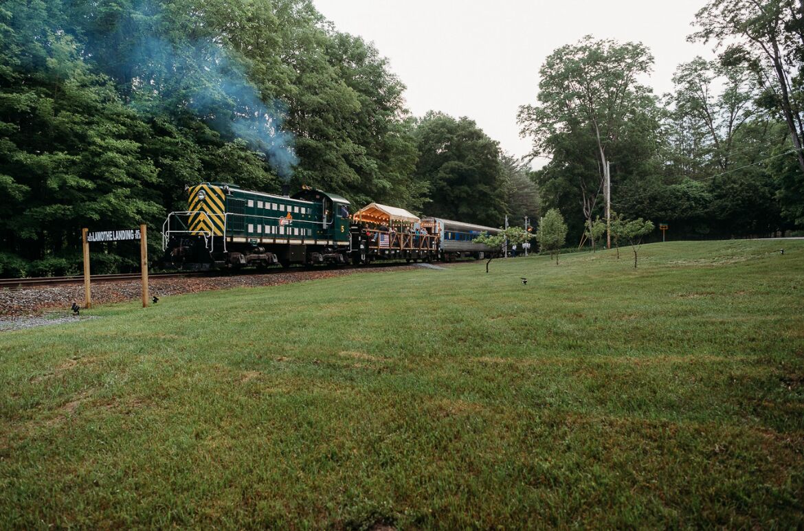Un train avec une locomotive verte et jaune et des wagons de voyageurs traverse une zone rurale, avec des champs couverts d’herbe et des arbres.