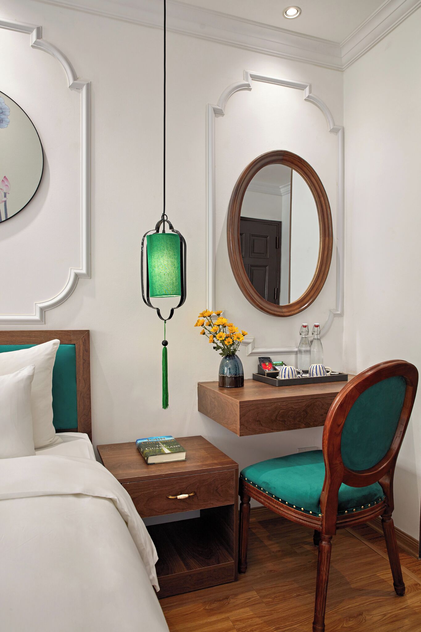A neatly arranged hotel room with a bed, a bedside table with a book, a chair, a desk with a mirror, and a hanging green lantern.