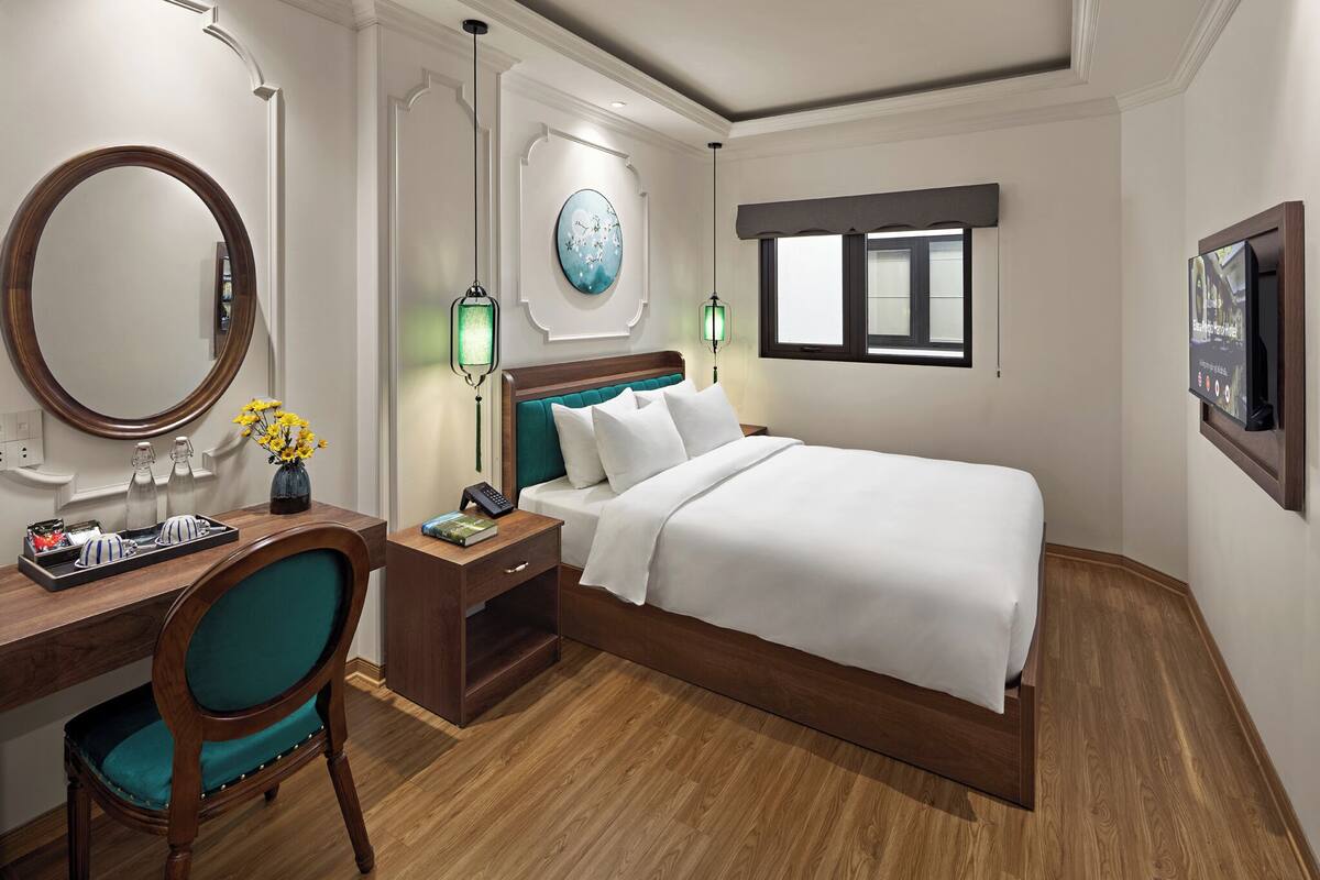 Une chambre d’hôtel avec un lit en bois, un bureau avec une chaise, une table de chevet, un miroir et un téléviseur fixé au mur.