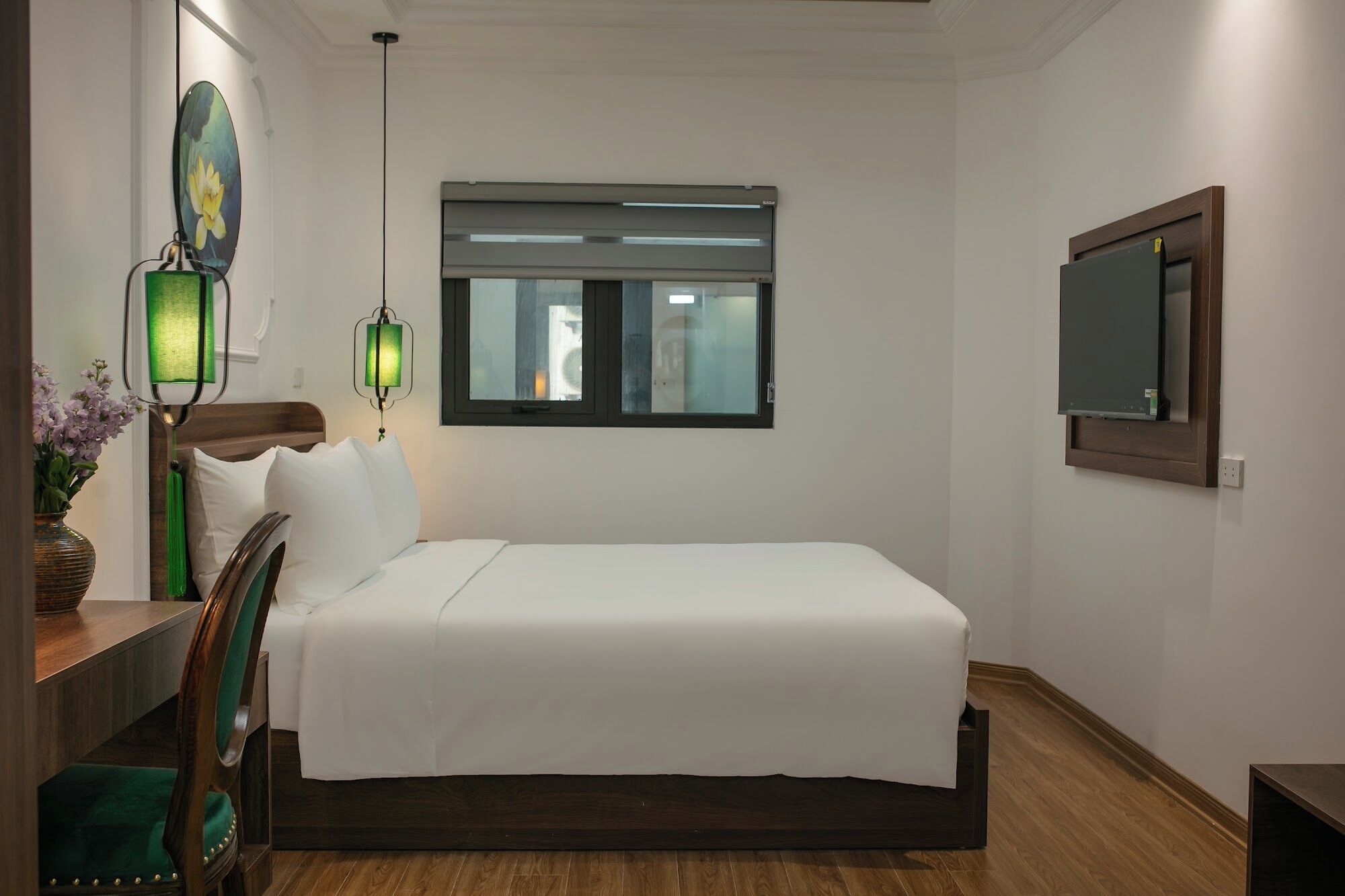 A hotel room with a bed, a desk with a chair, a window with blinds, a wall-mounted TV, and two wall sconces.