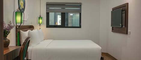 A hotel room with a bed, a desk with a chair, a window with blinds, a wall-mounted TV, and two wall sconces.