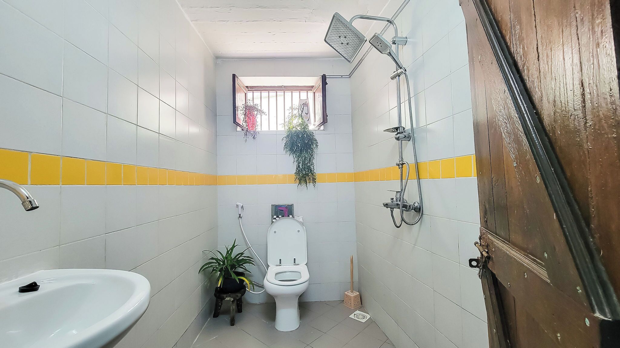 A small bathroom with white and yellow tiles, a toilet, a sink, a showerhead, and a window with a plant.