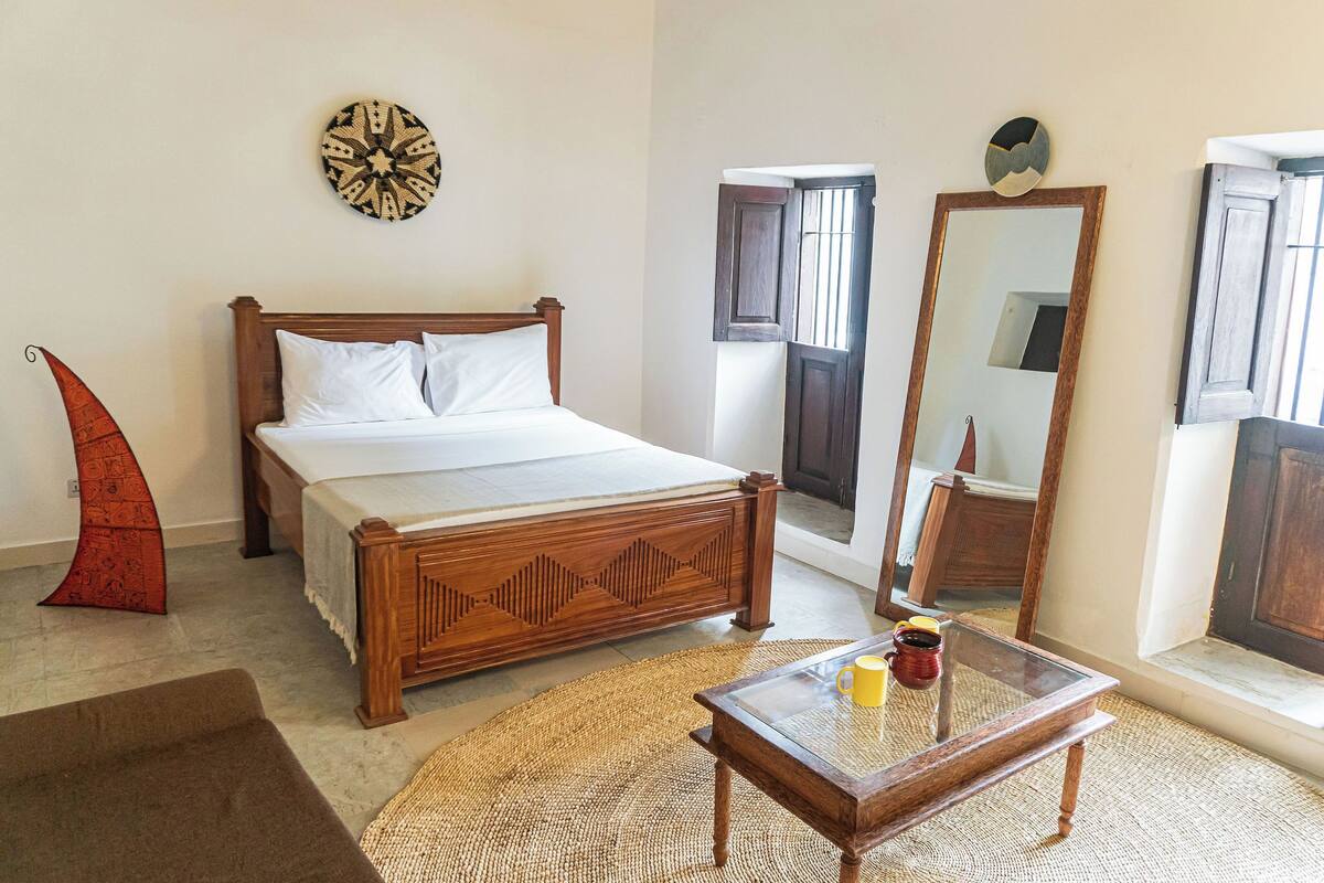 Un dormitorio con una cama de madera, una mesa de centro de vidrio, un tapete tejido y un espejo montado en la pared.