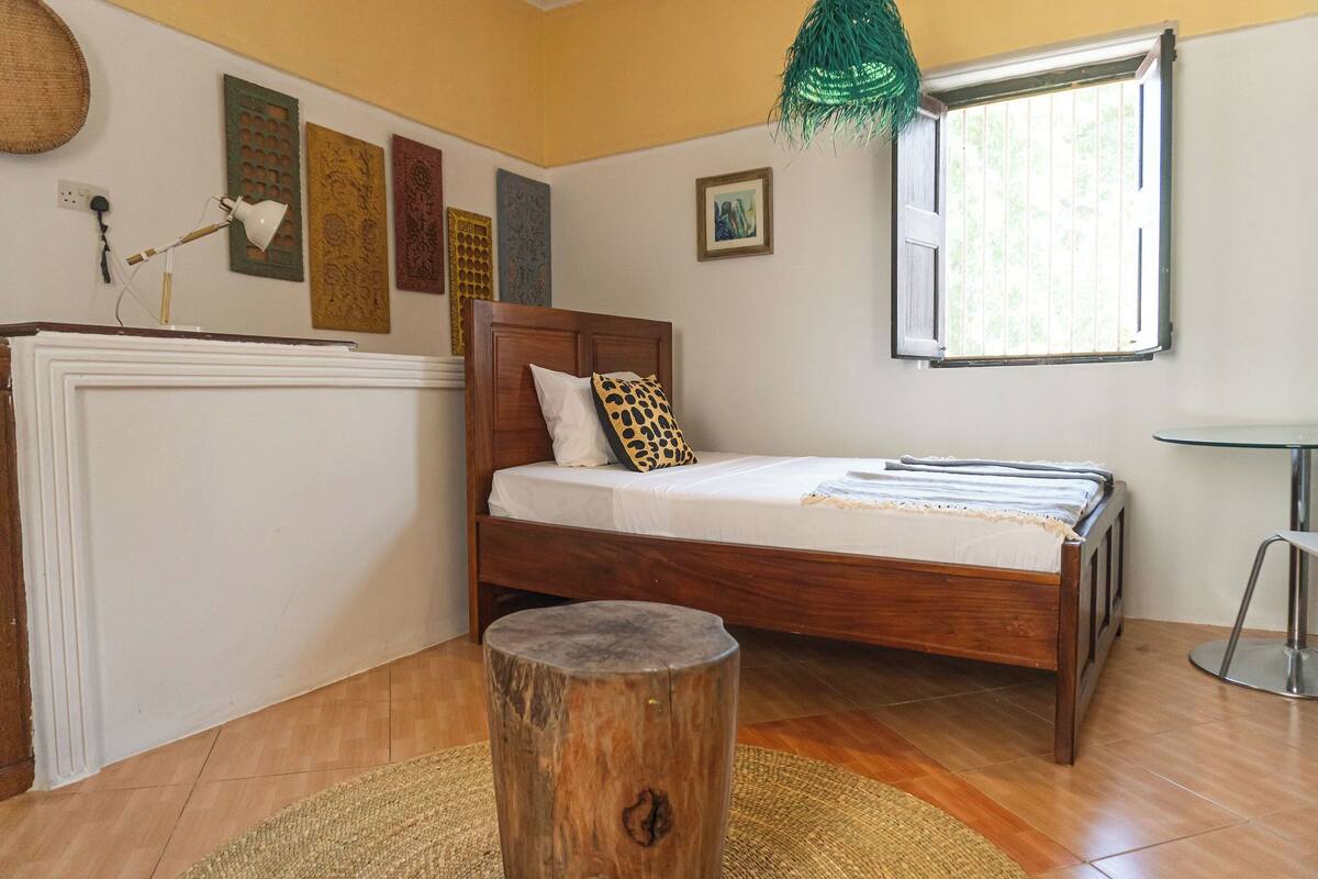 Un dormitorio con cama de madera, un taburete pequeño y redondo, una ventana con vistas y decoración en la pared.
