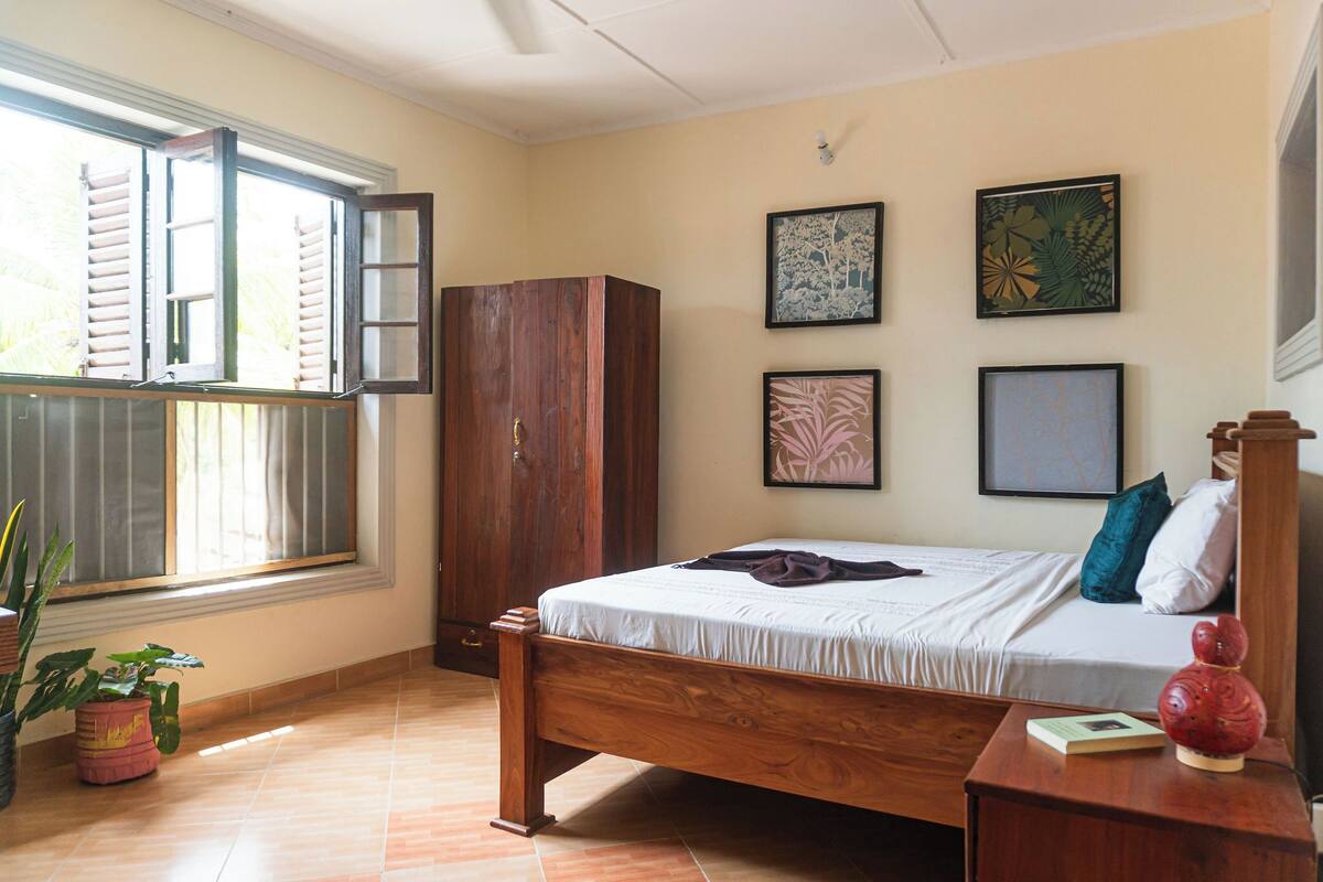 Un dormitorio con una cama de madera, un armario y una ventana con contraventanas. Hay cuatro obras de arte enmarcadas en la pared.