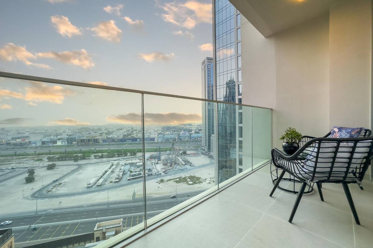 A balcony with a glass railing overlooking a cityscape with high-rise buildings and a highway. There is a chair and a small potted plant on the balcony.