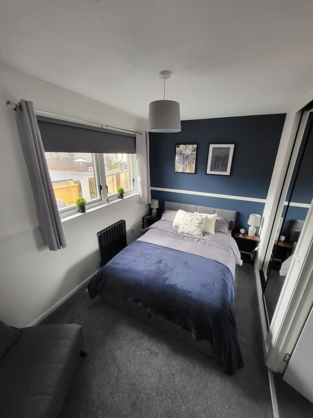 A bedroom with a bed, bedside tables, a radiator, a window with curtains, and artwork on the wall.