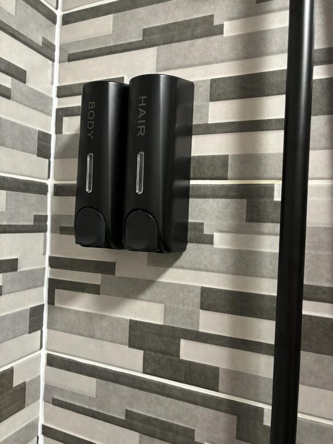 Ein Badezimmer im modernen Stil, mit einer Wand mit Muster und zwei schwarzen Spendern, auf denen „BODY“ und „HAIR“ stehen.