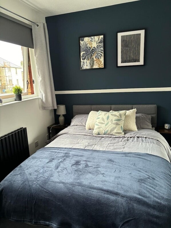 Un dormitorio con cama, dos obras de arte en la pared, una ventana con cortinas, un radiador y una mesita de noche con lámpara.