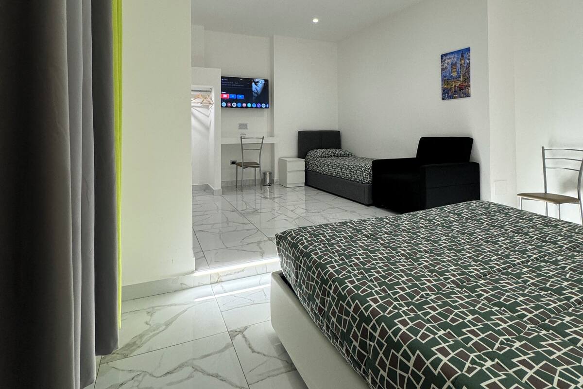 A modern hotel room with a large bed, a desk, and a chair. There is a TV mounted on the wall and a painting above the desk.