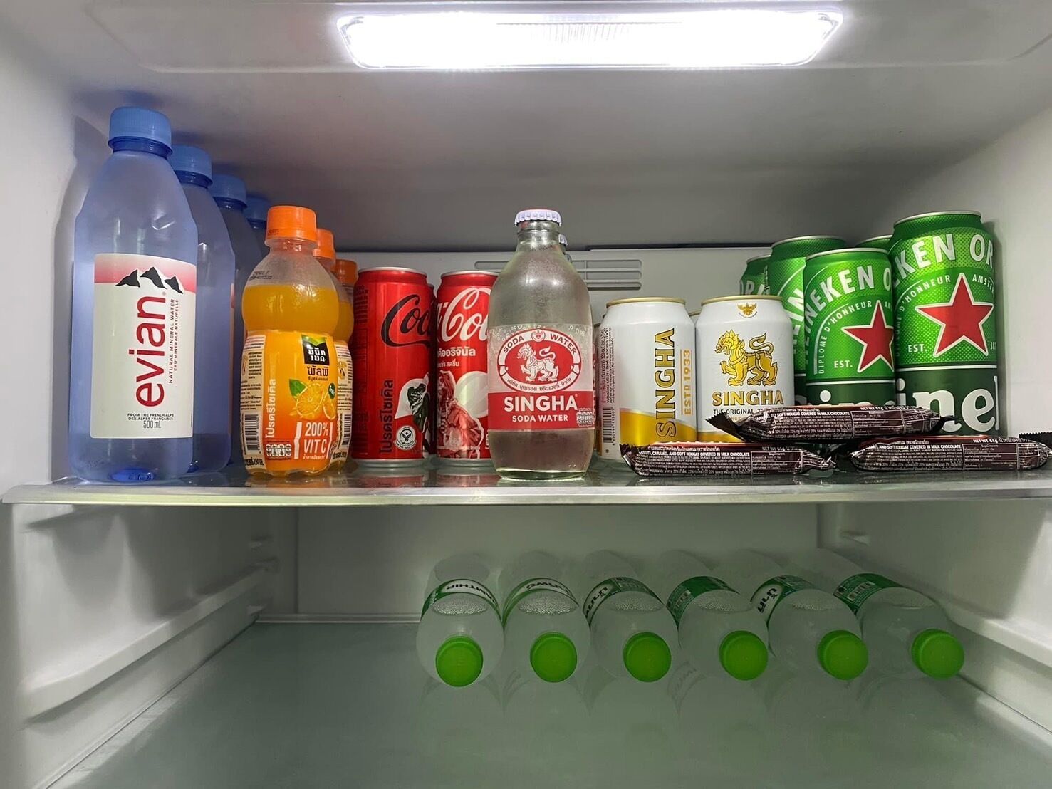 A refrigerator stocked with beverages including Evian water, orange juice, Coca-Cola, Singha soda, and Heineken beer.
