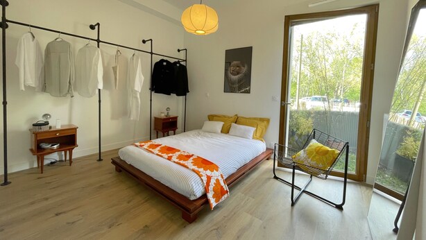 A bedroom with a bed, a chair, a hanging clothes rack, and a window with a view of trees and cars. - Immediate proximity to the Bordeaux hyper center (Bruges)