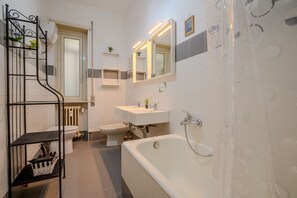 A bathroom with a bathtub, toilet, bidet, and a sink with a mirror above. There is a shelf with toiletries and a basket.
