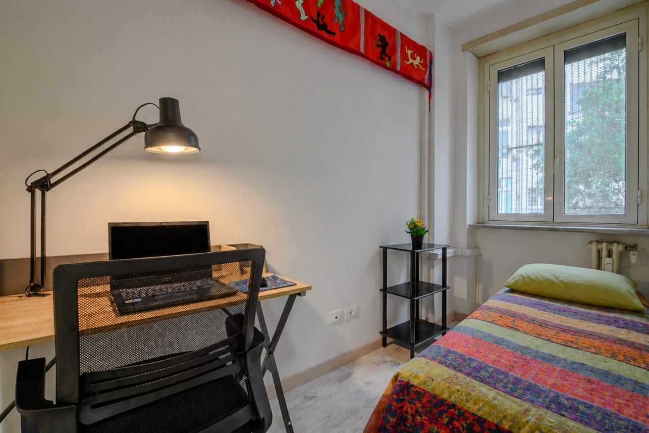 Une chambre à coucher avec un lit, un bureau avec un ordinateur, une chaise, une lampe, une fenêtre et un objet décoratif rouge accroché au mur.