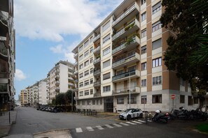 A multi-story residential building with balconies, parked cars, and a clear sky.