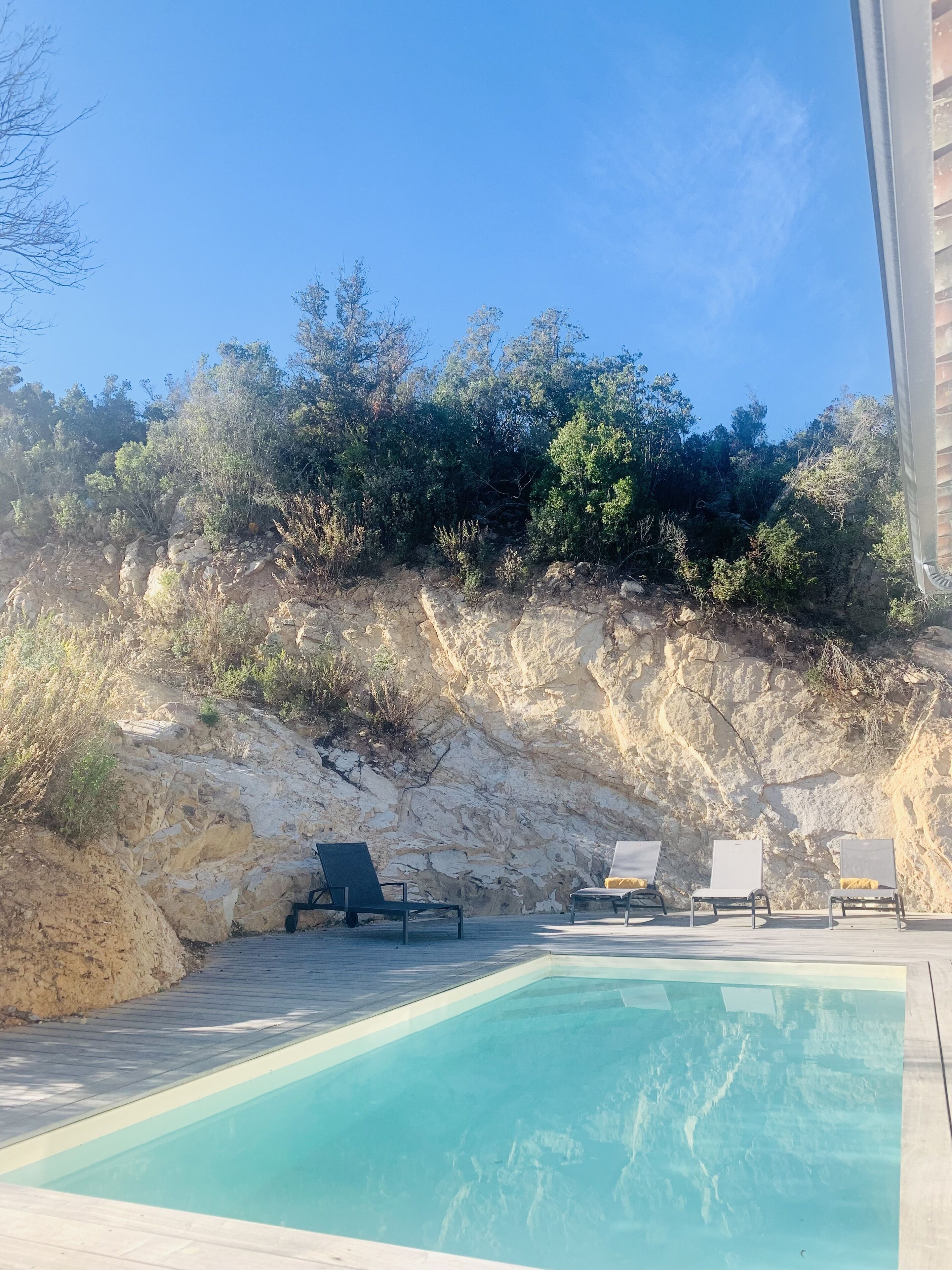 En swimmingpool med solstolar, omgiven av ett stenigt landskap och vegetation.