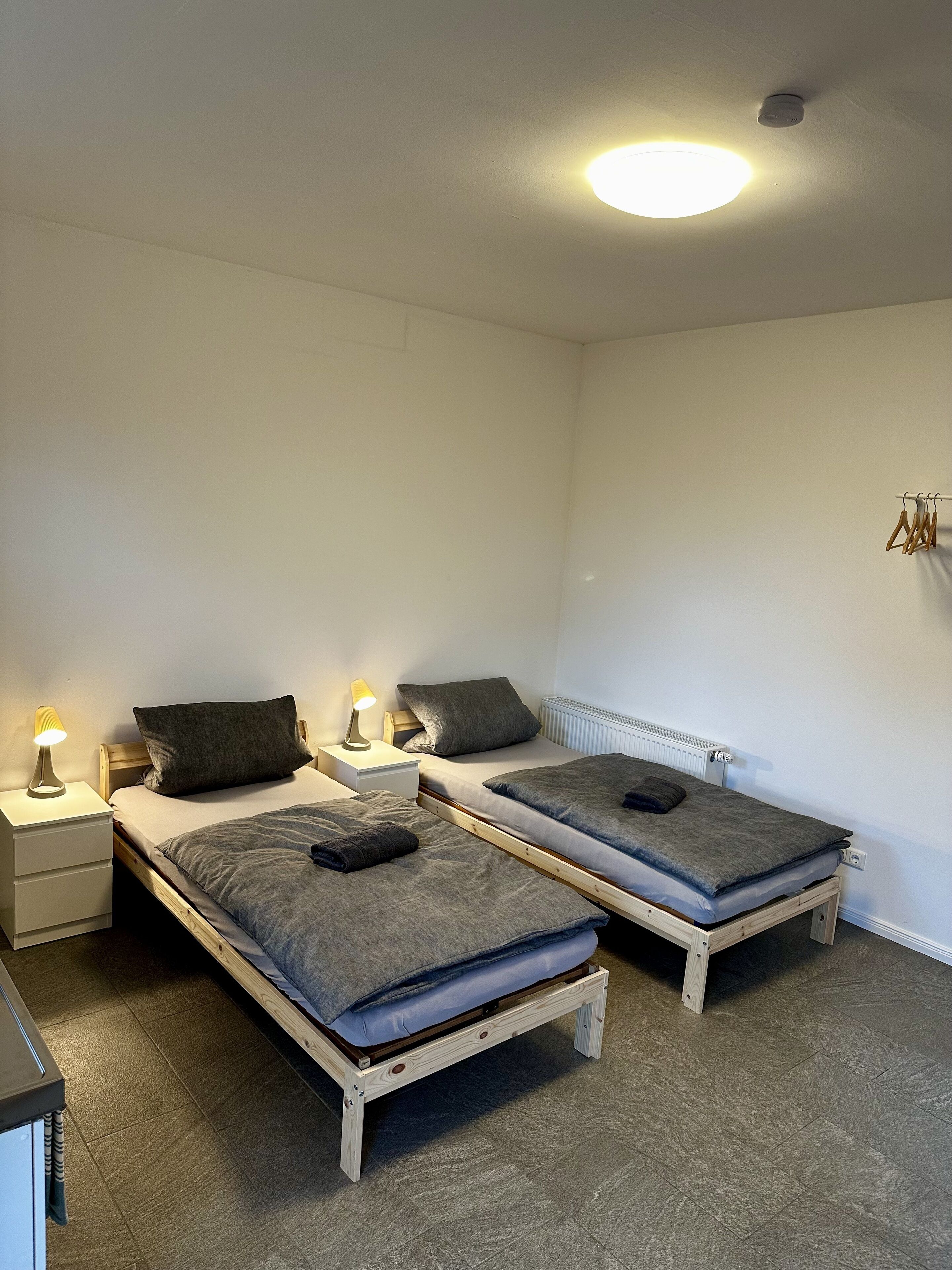 Une petite chambre propre, avec deux lits en bois, une table de chevet avec une lampe et un radiateur.