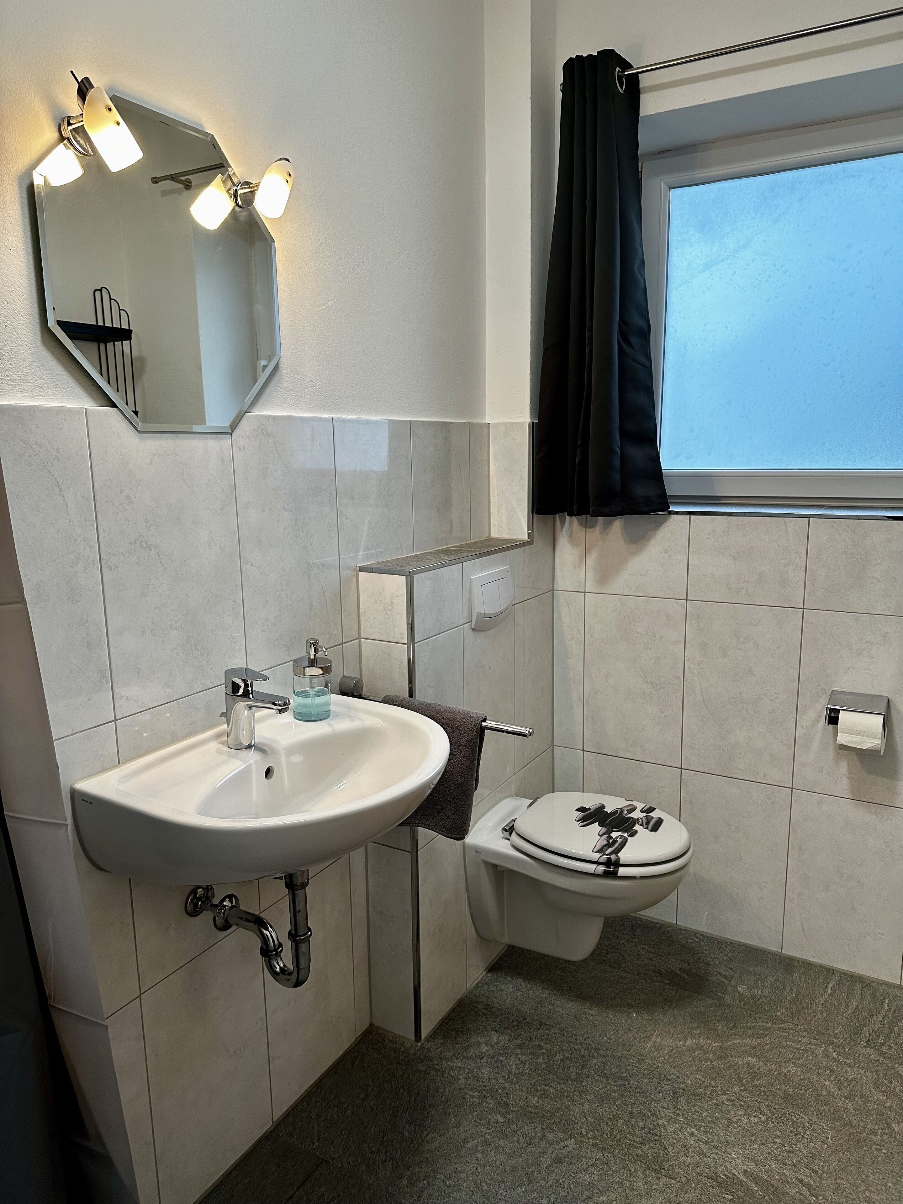 Ein Badezimmer mit einem weißen Waschbecken, einem Spiegel, einer Toilette und einem Fenster mit Vorhang.