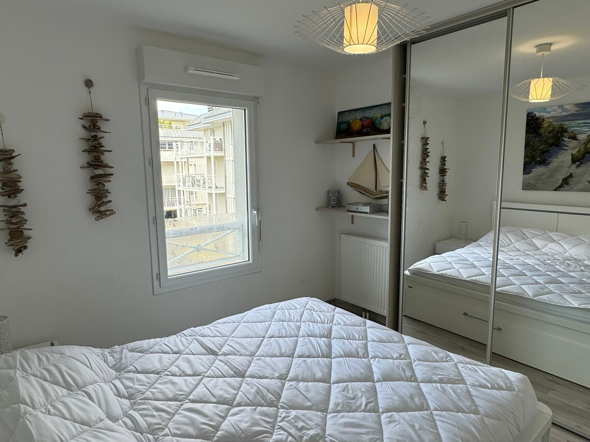 Ein Schlafzimmer mit Bett, Fenster, Heizkörper, einem Regal mit Gegenständen und einem Spiegel.
