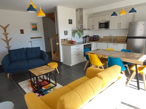 A modern kitchen with white cabinets, a stainless steel refrigerator, and a microwave. There is a dining table with yellow chairs and a blue sofa in the living area. - New home 2024, modern decor, near beach, free bikes, pets allowed (Saint-Jean-de-Monts)