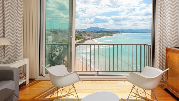 Un balcon donnant sur une plage et l’océan, aménagé avec deux chaises blanches et une petite table.