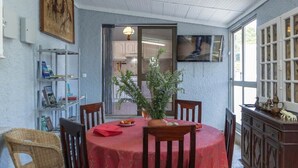 Un comedor con una mesa redonda cubierta por un mantel rojo, rodeada de sillas de madera. Hay una planta en maceta sobre la mesa y un televisor montado en la pared.