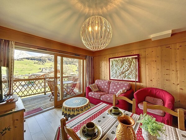 Um quarto aconchegante com decoração em madeira, um sofá vermelho, uma mesa de jantar com toalha de mesa estampada e vista para uma paisagem montanhosa através de grandes janelas.