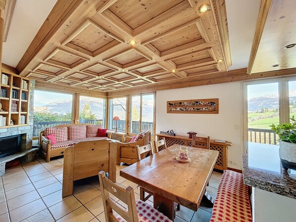 A cozy room with a wooden ceiling, a fireplace, a checkered sofa, a dining table, and a view of mountains through large windows.