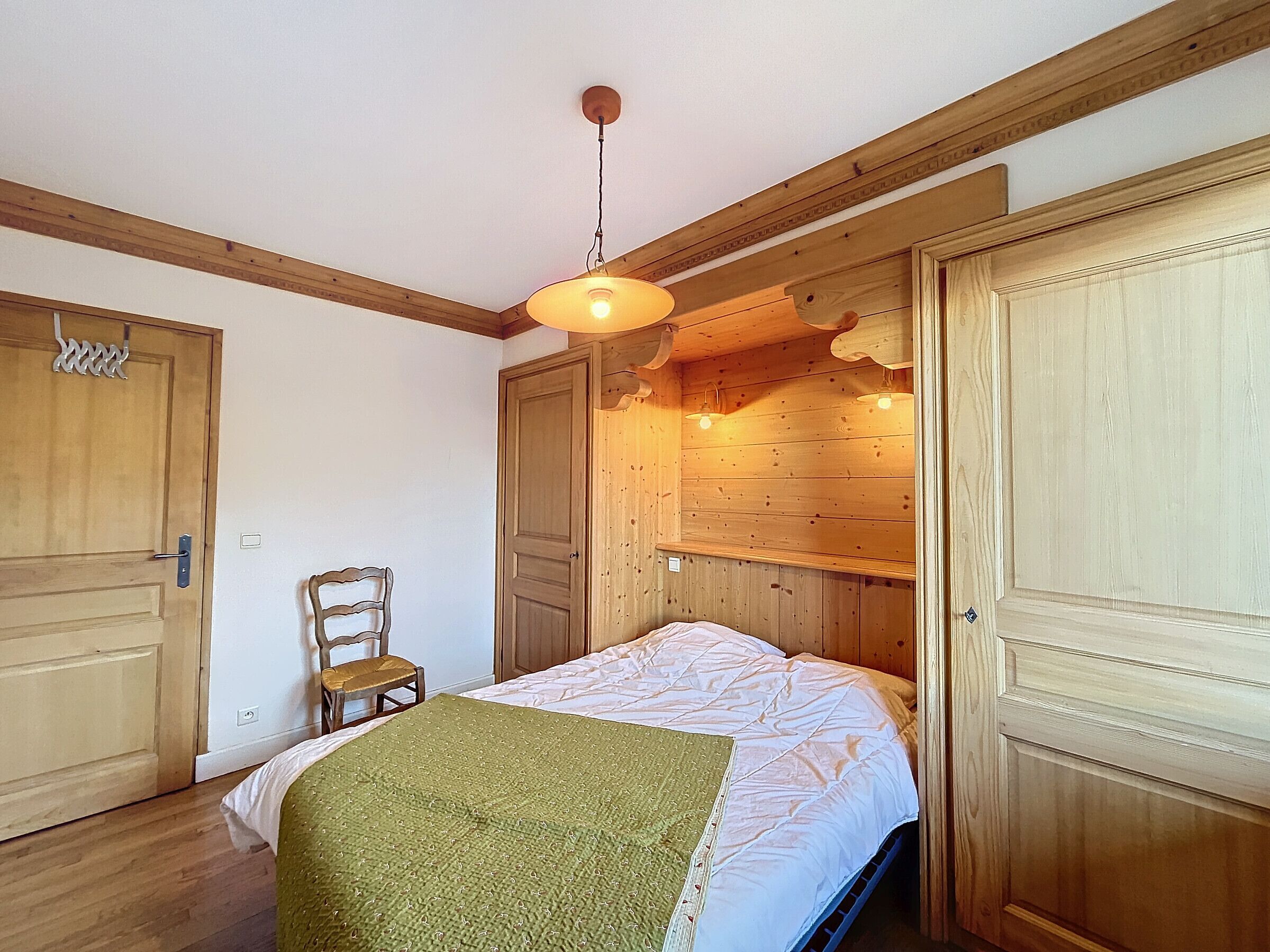 A bedroom with wooden walls, a bed with a green blanket, a wooden chair, and a hanging light fixture.