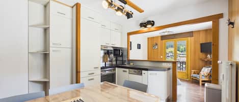A modern kitchen with white cabinetry, a wooden dining table, and a view of a balcony with a chair.
