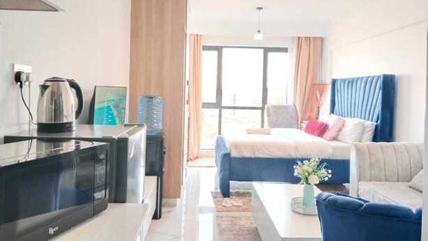A modern hotel room with a kitchenette, a bed with blue and white bedding, a blue armchair, and a view of the outdoors. - Comfort Homestay Kilimani (Nairobi)