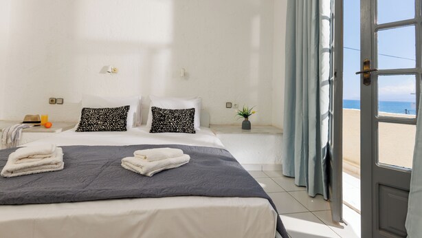 A hotel room with a bed, a window with curtains, a bedside table, and a view of the beach. - Kings Hotel by Estia (Hersonissos)